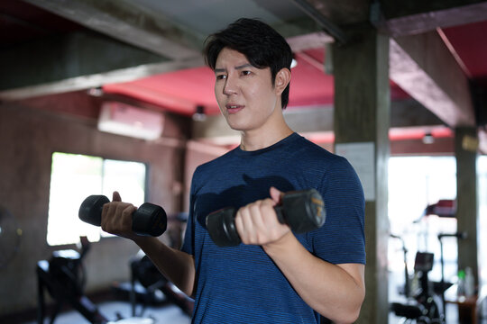 Young Asian man performing dumbbell bicep curls during strength training session at modern fitness gym