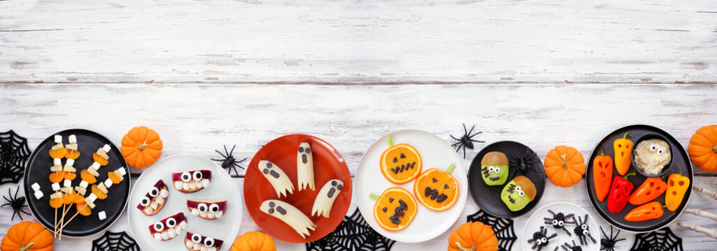 Healthy Halloween fruit and vegetable snacks. Variety of fun, spooky treats. Top down view bottom border over a white wood banner background with copy space.