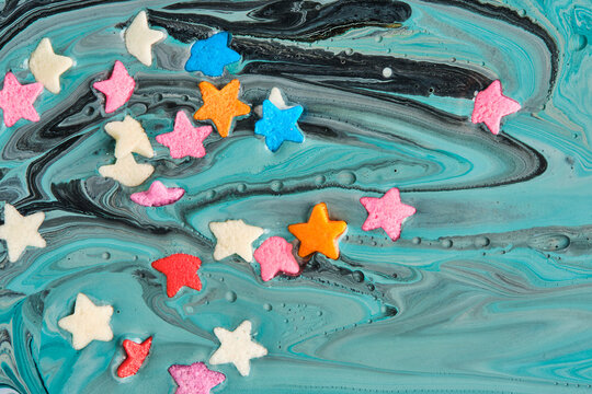 Colorful star shaped sprinkles floating on top dark turquoise liquid paint swirl.