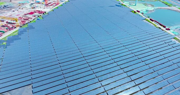 An aerial view of an expansive photovoltaic solar panel farm located next to an industrial construction area and water under bright sunlight.