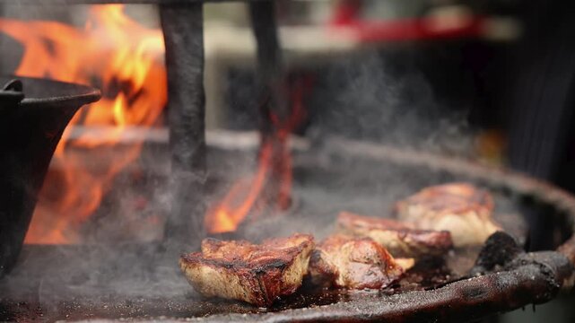 Meat steaks grilling on open fire with smoke and flame. Barbecue cooking on charcoal grill at street food market. Roasted pork preparing on hot metal plate, delicious street food concept.