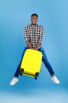 Happy Smiling Black Male Traveler Jumping With Yellow Suitcase In Bright Studio Setting, Showing Excitement For Upcoming Trip. Ready For Adventure And Fun, Vacation Concept.