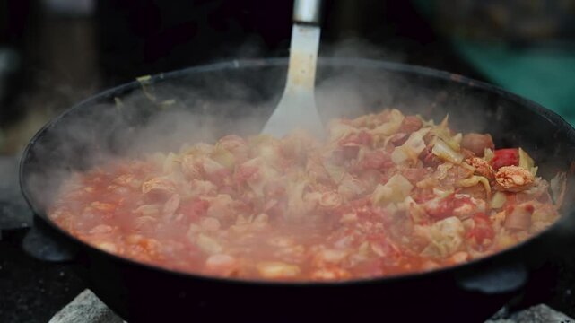 Cooking vegetable stew in large pan outdoors. Steam rising from tomato and cabbage dish. Street food preparation with spatula, traditional cuisine concept