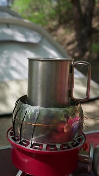 A stainless steel cup sits on top of a portable gas stove with a metal wind shield. The camping burner is set up outdoors on a sunny day at a campsite. A warm beverage is prepared in nature.