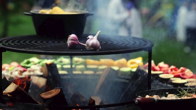 Garlic and vegetables grilling on open fire at outdoor barbecue. Firewood burning under grill grate with smoke. Street food cooking on fire concept