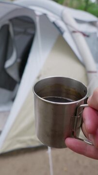 A hand holds a stainless steel mug with hot coffee in front of a camping tent set up in the forest. Concept of morning energy, outdoor relaxation, and peaceful camping retreat.