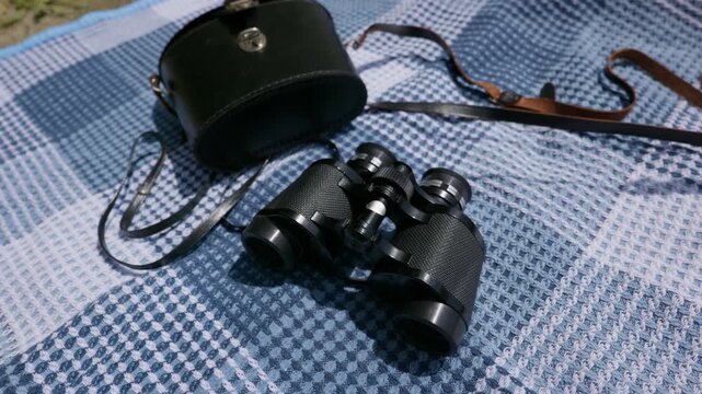 Vintage black binoculars and a leather carrying case lie on a blue plaid picnic blanket in the sun. Concept of outdoor exploration, sight-seeing, nature observation, and adventure.