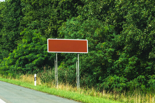 Blank brown tourist sign mounted beside a road with dense green foliage in the background, concept of road signage mockup with copy space