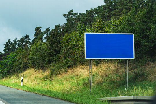 Blank blue motorway sign beside a grassy roadside with green trees under an overcast sky, concept of road signage mockup with copy space