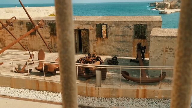 Historic rusted anchors and heavy naval chains displayed at Fort St Elmo along the coastline of Valletta, Malta, overlooking the turquoise Mediterranean Sea and harbor fortifications on a sunny day.

