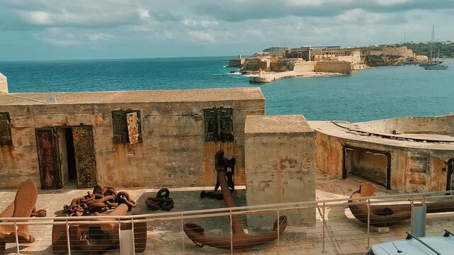 Historic rusted anchors and heavy naval chains displayed at Fort St Elmo along the coastline of Valletta, Malta, overlooking the turquoise Mediterranean Sea and harbor fortifications on a sunny day.

