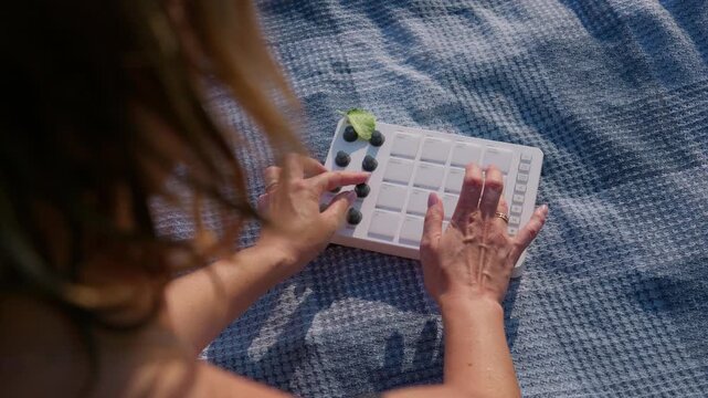 A musician turns rotary knobs on a portable MIDI controller while relaxing outdoors on a woven blanket. Concept of sound design workflow, outdoor beatmaking, and creative sound synthesis.