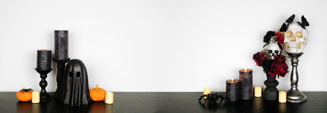 Halloween gothic shelf decor with copy space against a white wall banner background. Black shelf with glowing candles, dark flowers, skulls, pumpkins, ghost and spiders.