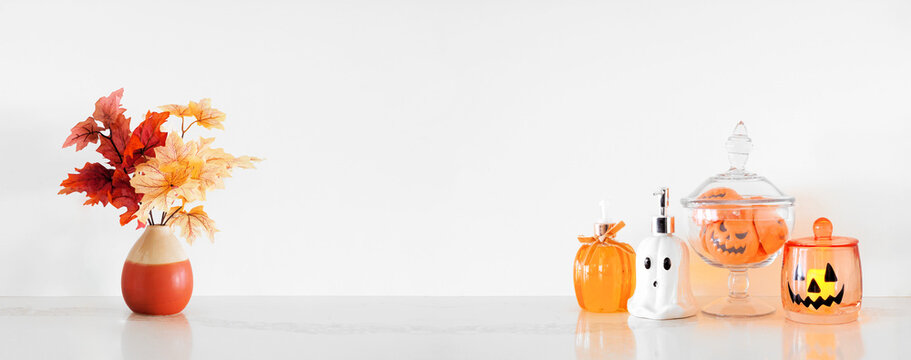Halloween pumpkin and ghost theme body care on a white stone countertop. Banner against a white wall background with copy space. Bathroom decor concept. Soaps, lotion, bath fizzers.
