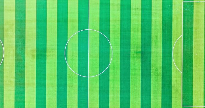 Aerial view of a green soccer pitch featuring alternating vertical stripes and white field lines under bright natural sunlight.