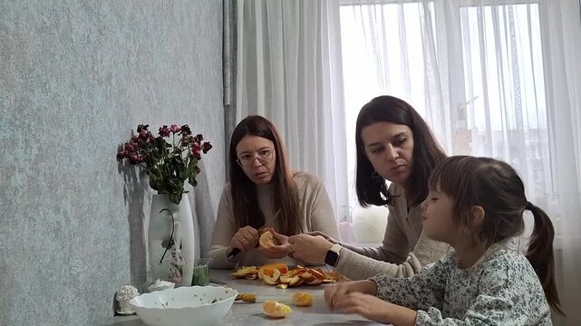 Women and a girl sitting at the table peeling and eating oranges in a bright room with plants and flowers