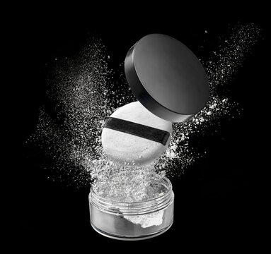 Loose powder compact with open lid and makeup puff floating above sparkling white particles on black studio background
