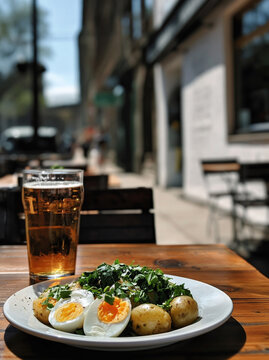 Sałatka z ugotowanymi ziemniakami, jajkami na p&oacute;łtwardo i dużą ilością zielonych zi&oacute;ł na talerzu oraz szklanka piwa na drewnianym stoliku w ogr&oacute;dku restauracyjnym.