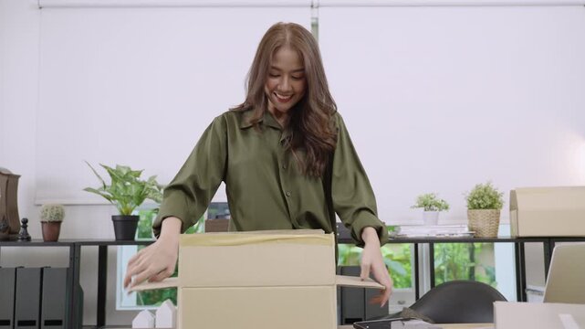 Shopping online concept. Happy attractive young Asian woman taking open box and checking shirt with long sleeved cream color in her online shopping order with smiling and cheerful in her home office. 