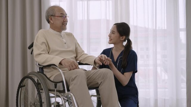 Beautiful young Asian woman nurse caregiver in uniform advises, encourages, and take care of a senior man patient paralyzed with a problem at legs while sitting in a wheelchair in a hospital room. 