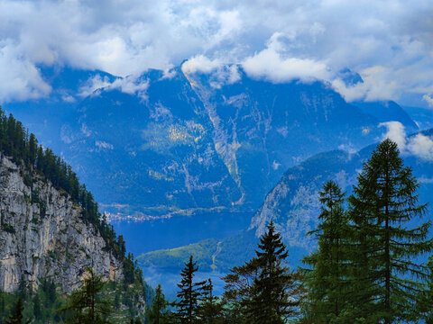 Majestic blue mountains rise under fluffy clouds, surrounded by lush evergreen trees and a tranquil lake in the valley.
