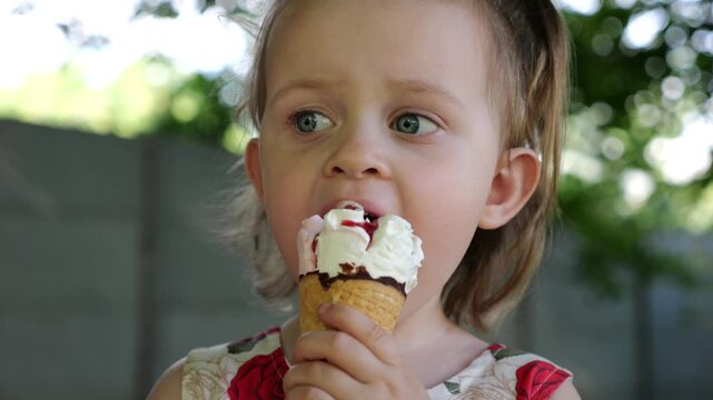 Portrait of attractive small girl eating delicious ice cream in a waffle cone outdoor. Beautiful child biting cold sweet dessert in summer garden. Cute kid enjoying tasty refreshing food on sunny day