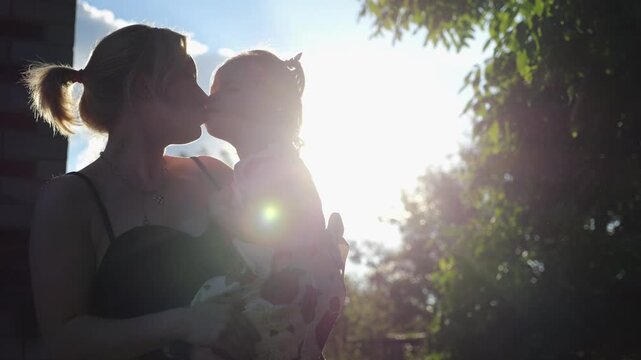 Beautiful silhouette of young mom kiss her cute small girl in summer garden. Happy parent and child sharing sweet emotional moment in nature. Motherhood, parental care and unconditional love concept