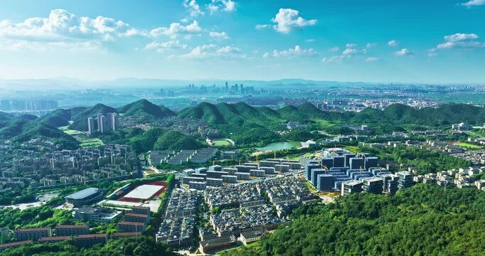 Aerial panorama shows urban buildings nestled among green hills under bright sunlight, with residential blocks and a distant skyline beneath a cloudy sky.