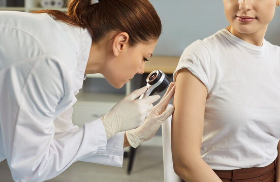 Professional dermatologist examining mole on female patients arm using handheld dermatoscope during visit at hospital or clinic. Mole check-up, melanoma awareness, skin cancer prevention concept