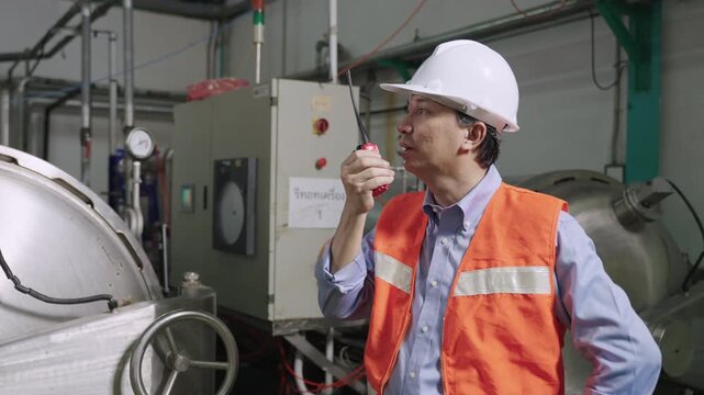 Professional Asian man maintenance engineer in uniform of the factory with a white safety hardhat standing using a walkie talkie radio for talking and discussing communication in the factory industry.