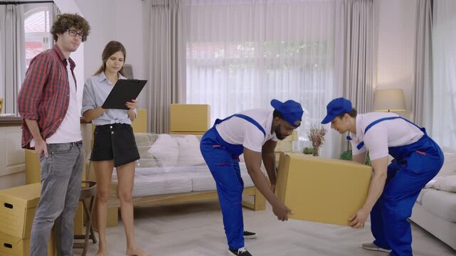 Team professional movers in uniform are moving furniture and cardboard boxes during a home relocation, while a young couple of homeowners, holding a clipboard, are checking for delivery at their home.