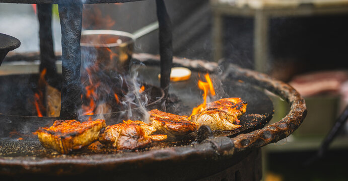 Meat steaks grilling on open fire with smoke and flame. Barbecue cooking on charcoal grill at street food market. Roasted pork preparing on hot metal plate, delicious street food concept.