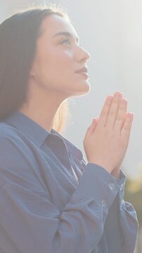Portrait of young woman praying with closed eyes to God asking for blessing, help, forgiveness outdoors. Pretty lady clasping hands wishing luck in sunny urban city street. Town religion lifestyles