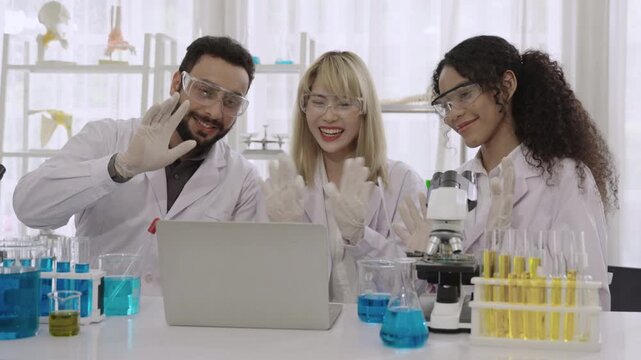 Group of happy men and women ethnic diversity, scientists and researchers wearing lab coats and goggles sitting and using a computer laptop for an online conference meeting together in the laboratory.