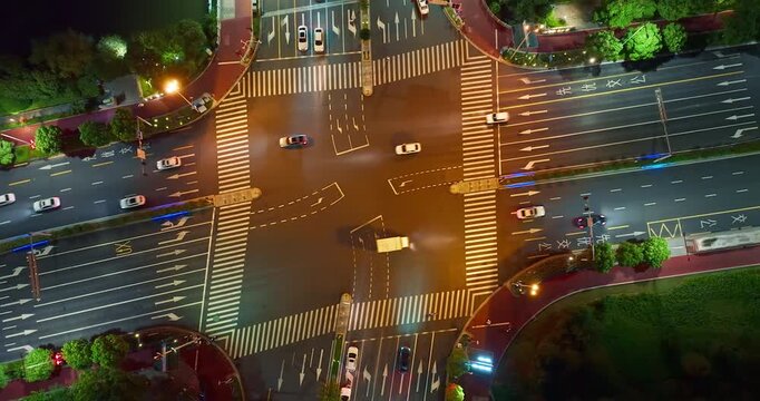 Aerial view of a city road intersection at night, showing illuminated asphalt streets, pedestrian crosswalks, moving cars, and warm streetlight beams.