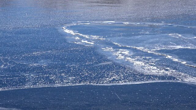 Cracked lake ice covers a cold basin, forming broad fracture lines across the frozen surface. Broken sheets and solid winter crust create a rugged panorama with frigid seasonal texture.