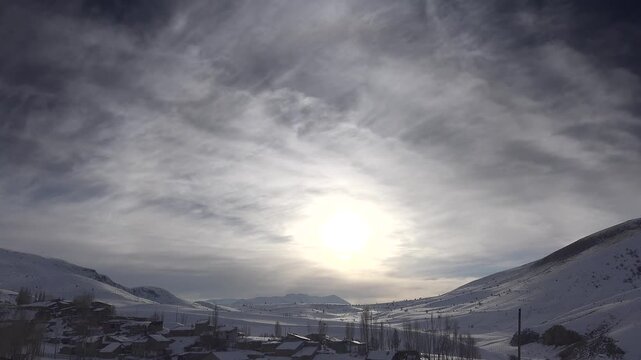 Traditional homes dot a snowy valley beneath cirrus clouds, creating a quiet highland scene. Rural settlement spreads across cold hills with mountain houses, winter light, and open sky.