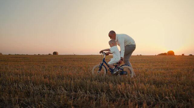 Father teaching young son to ride a bicycle across a golden harvested field at sunset, sharing support, encouragement, and joyful bonding in peaceful countryside