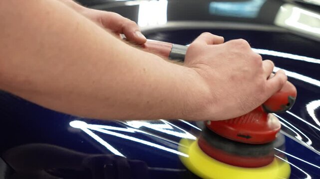 Professional car detailer worker polishes a luxury vehicle hood paint using an electric dual action orbital polishing machine tool with a yellow foam pad buffer in a garage workshop service.