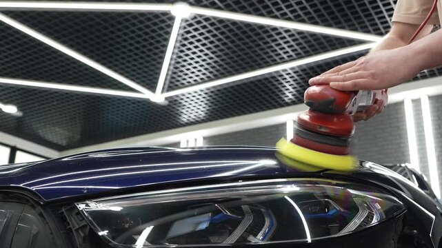 Professional car detailer worker polishes a luxury vehicle hood paint using an electric dual action orbital polishing machine tool with a yellow foam pad buffer in a garage workshop service.