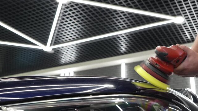 Professional car detailer worker polishes a luxury vehicle hood paint using an electric dual action orbital polishing machine tool with a yellow foam pad buffer in a garage workshop service.