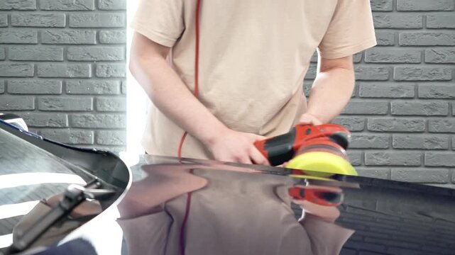 Professional car detailer worker polishes a luxury vehicle hood paint using an electric dual action orbital polishing machine tool with a yellow foam pad buffer in a garage workshop service.