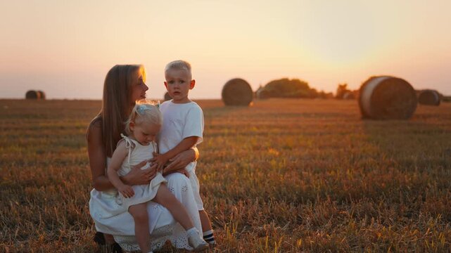 Loving mother embracing her young children in a golden harvested field at sunset, enjoying peaceful family time together in warm summer countryside light