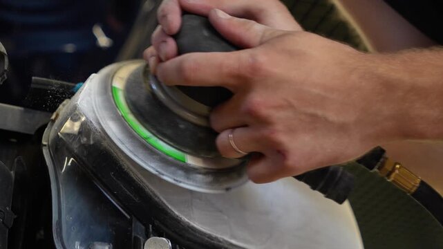 Car detailing technician sanding foggy plastic headlight close up, man hands using orbital sander machine with sandpaper for automotive headlamp restoration and scratch repair. 