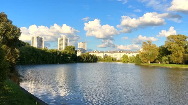 Lake in city park with trees in Autumn landscape. Tall buildings rise background. Buildings near lake under blue sky with clouds in afternoon. Residential condo buildings at lake surrounded by trees. 