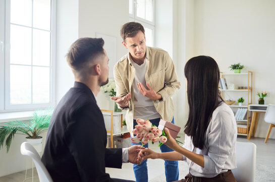 Jealous man expressing anger while witnessing romantic proposal as woman holding flowers and gift. Intense love triangle filled with conflict, tension, betrayal and dramatic emotions indoors.