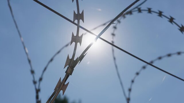 A close-up view shows sharp razor wire blades shining against a bright midday sun in clear blue sky. Concept of perimeter security, border protection, and industrial defense.