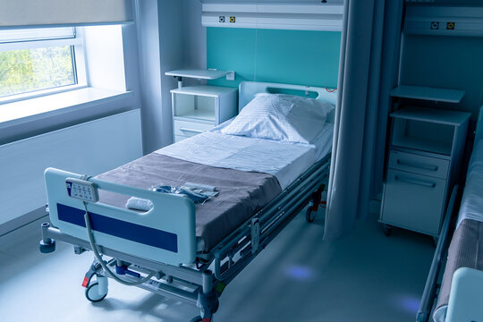 A made hospital bed stands by the window in a quiet patient room. A bedside cabinet and an overbed table wait next to it. Comfortable ward for recovery in a new modern hospital