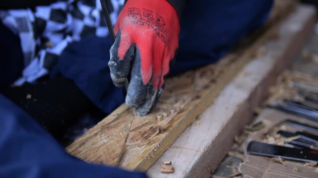 Gloved hand carving wood across narrow plank with steady chisel work. Raised ornament and fresh shavings show careful artisan detail, with rough board and tools framing focused manual craftsmanship.