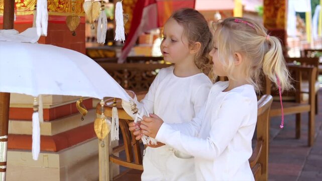 Two young girls hold ornate white parasol during ceremony ritual in temple setting. Their white clothing and careful hands suggest quiet participation as they stand beside decorated posts and tassels.